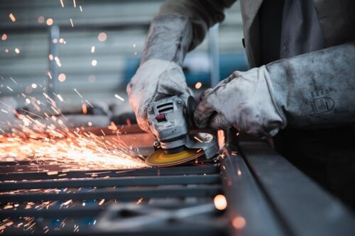 Nahaufnahme eines Arbeiters mit Schutzhandschuhen, der mit einem Winkelschleifer Metall bearbeitet, wobei ein intensiver Funkenregen entsteht