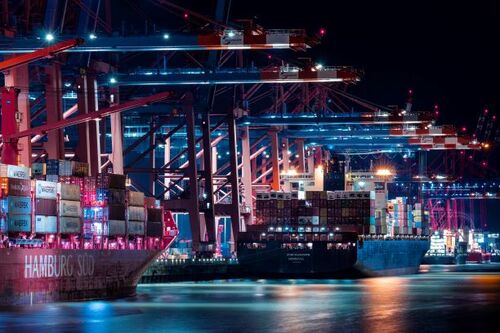 Ein nächtlich beleuchteter Containerhafen mit mehreren großen Frachtschiffen, die an Verladestationen mit riesigen Kränen liegen | Sabine Verheyen